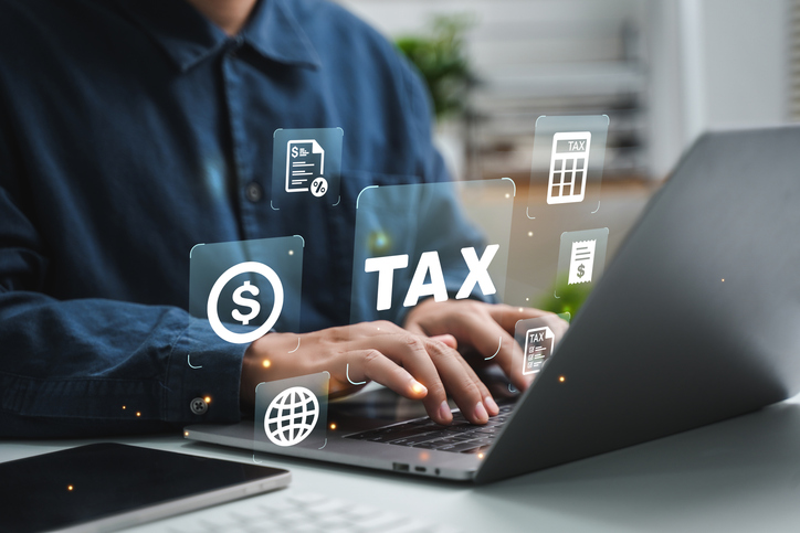Person typing on laptop with the word "tax" and floating icons of money, calculator, and invoices