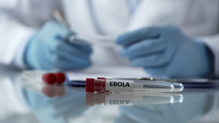 Lab worker in protective equipment taking notes beside a flask marked “Ebola.”