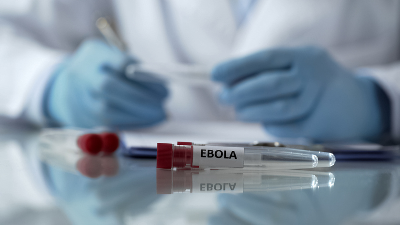 Lab worker in protective equipment taking notes beside a flask marked “Ebola.”
